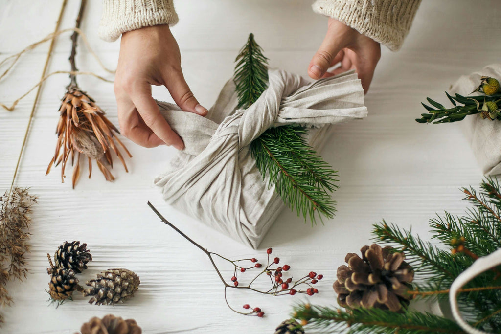 Person wrapping a gift with fabric and greenery, surrounded by pine cones and winter foliage.