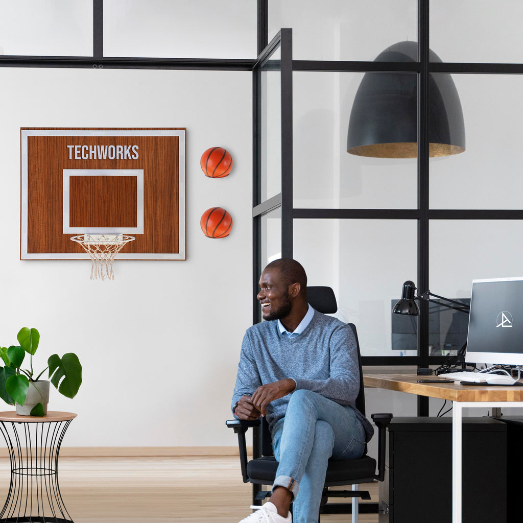 A man sitting in an office with a wooden wall basketball hoop and two basketballs, enhancing workspace entertainment.